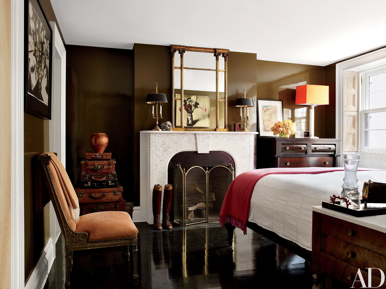 The intimate guest room, painted in Benjamin Moore’s Grizzly Bear Brown, features an Italian neoclassical slipper chair from JF Chen covered in orange suede; on the marble mantel is a pair of brass bouillotte lamps by Circa Lighting.