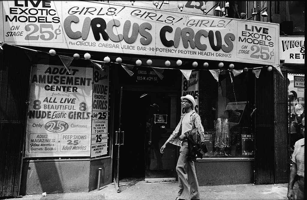 Exterior view of Circus Circus, an 'adult amusement center,' in Times Square as a man walks past wearing a hat and carrying his coat over his arm, New York, New York, 1970s. The marquee advertises exotic models, nude girls, books, magazines, games, and peep shows, as well as a juice bar and 'high-potency protein health drinks.' (Getty Images)